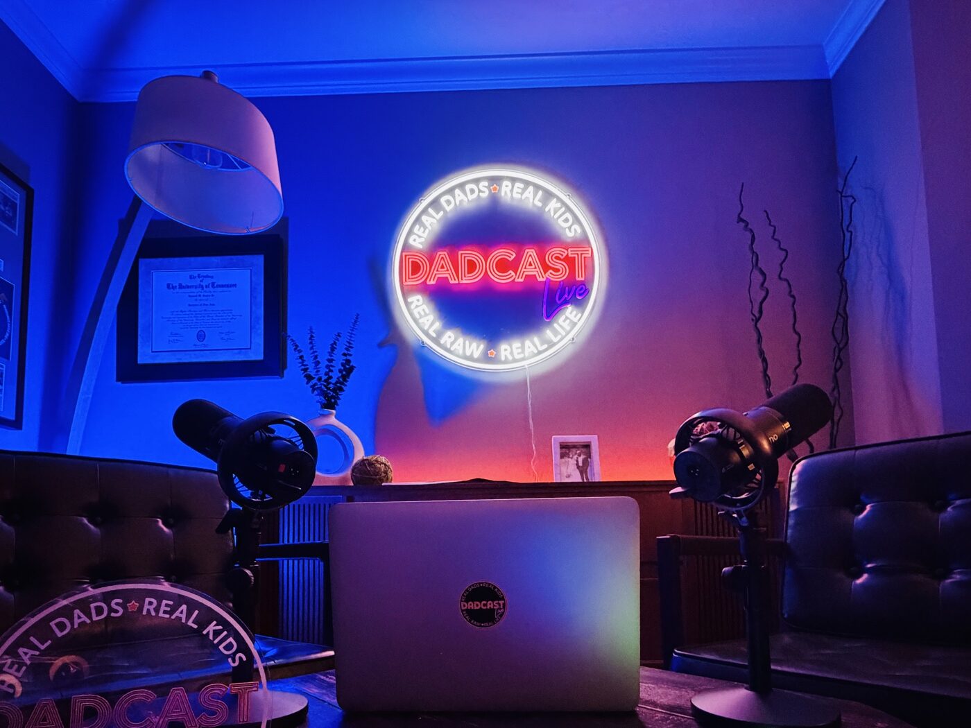 Close-up of the Dadcast Live neon sign glowing pink and blue with Rode microphones in the foreground