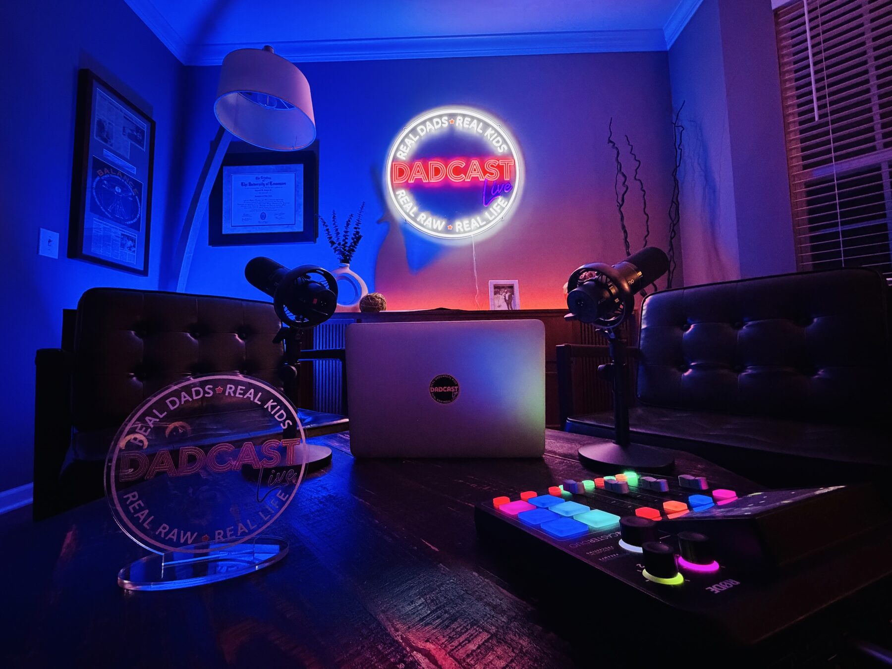 The Dadcast Live studio, with a neon sign that reads DADCAST live, Real Dads, Real Kids, Real Raw, Real Life, with Rode broadcast microphones and a Rodecaster board